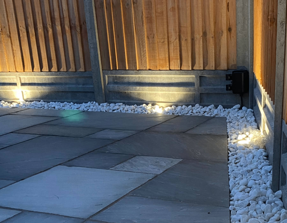 Professional outdoor lighting installation showcasing modern LED ground lights illuminating a contemporary wooden fence and paved patio area at dusk
