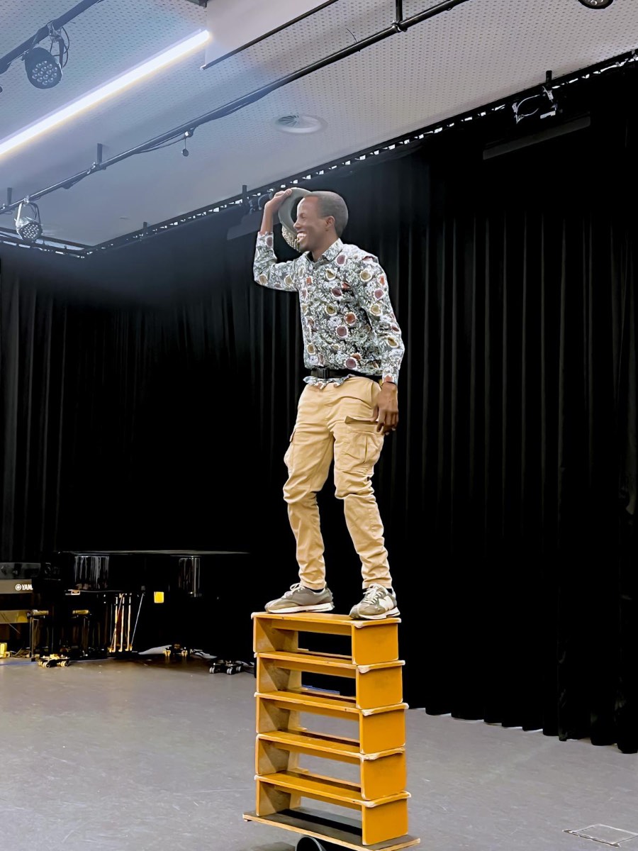 The King of Balance standing on stacked yellow crates performing balance act on stage with black curtain backdrop The King of Balance standing on stacked yellow crates performing balance act on stage with black curtain backdrop