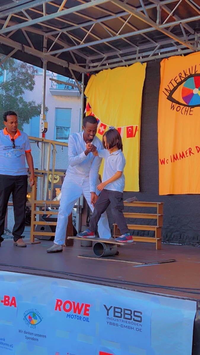 Kids Circus Workshop The King of Balance teaching a child rolla bolla balance skills on stage at Interkulturelle Woche event